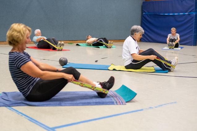 Senioren führen Rückenübungen mit Widerstandsbändern auf Matten in einer Sporthalle durch. Diese sind in verschiedenen Farben.