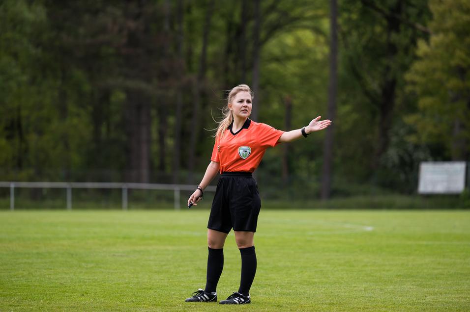 Schiedsrichterin in orangefarbenem Trikot zeigt in einem Fußballspiel gestikulierend auf dem grünen Spielfeld.