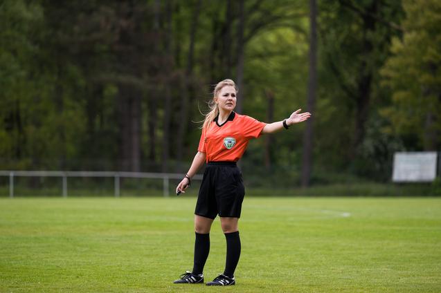 Schiedsrichterin in orangefarbenem Trikot zeigt in einem Fußballspiel gestikulierend auf dem grünen Spielfeld.
