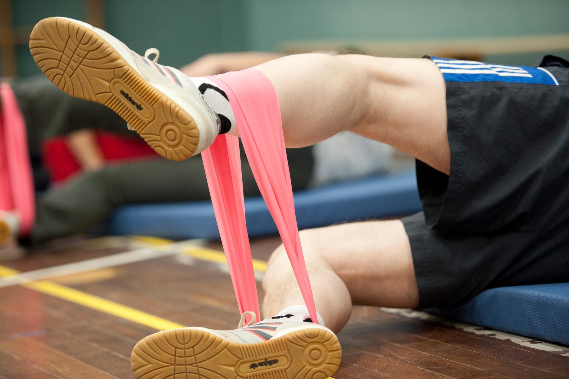 Bein eines Sportlers mit pinkem Widerstandsband, auf einer Sportmatte, im Hintergrund weitere Teilnehmer.