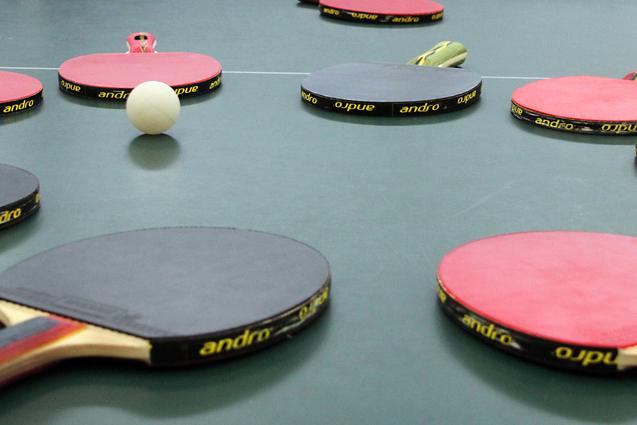 Mehrere Tischtennisschläger in verschiedenen Farben und ein Tischtennisball liegen auf einer grünen Tischtennisplatte.