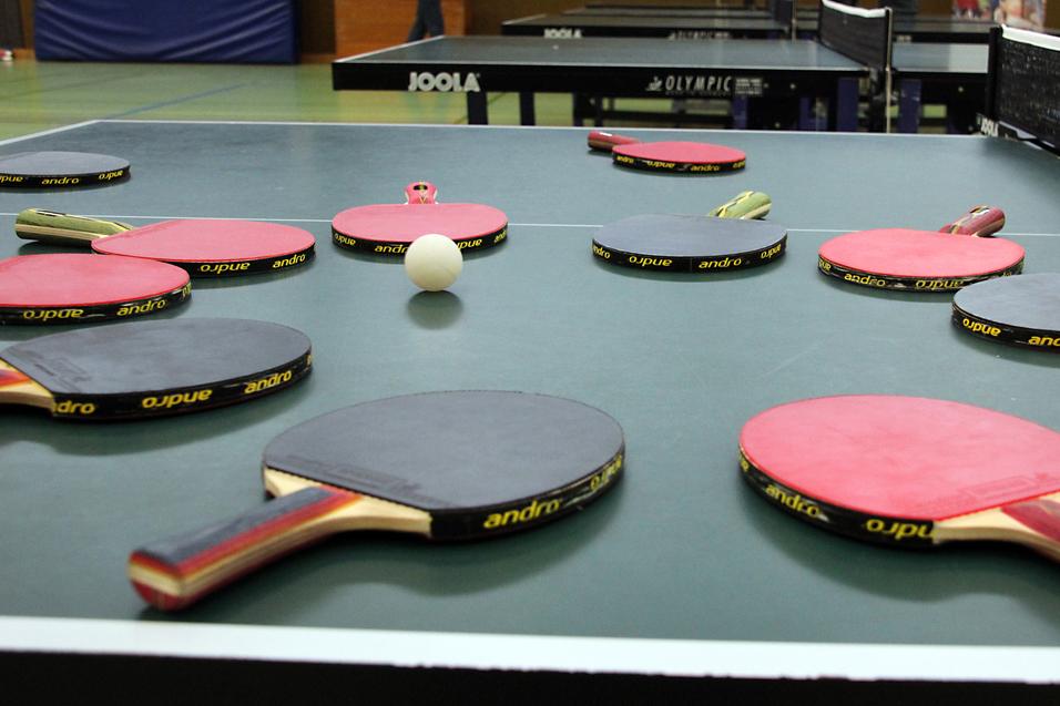 Mehrere Tischtennisschläger in verschiedenen Farben und ein Tischtennisball liegen auf einer grünen Tischtennisplatte.