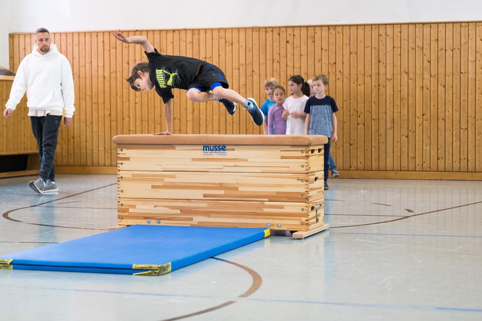 Ein Junge springt über ein Turngerät, während eine Gruppe Kinder und ein Trainer zuschauen. Sporthalle mit Holzverkleidung.