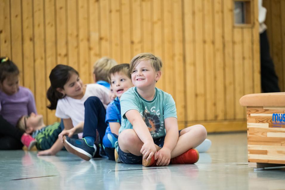 Gruppe von Kindern sitzt in einer Turnhalle, einige lachen, während ein Junge mit roten Schuhen lächelt.
